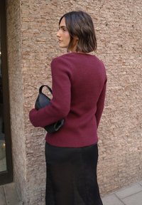 Bordowy dzianinowy sweter z okrągłym dekoltem i dopasowanym krojem, zestawiony z czarną skórzaną torbą oraz czarną półprzezroczystą spódnicą.