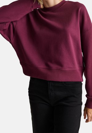 Femme portant un sweat-shirt bordeaux ample à manches longues et un pantalon noir, avec un bras levé pour toucher le cou.