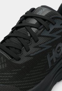 Μαύρο αθλητικό παπούτσι με υφασμάτινη υφή, δεμένα κορδόνια σε σταυρωτό σχέδιο και ζιγκ-ζαγκ μοτίβο στο πλάι κοντά σε λογότυπο "HOKA".