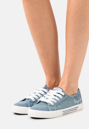 Baskets en toile bleue avec lacets blancs et semelle en caoutchouc. Présentent un logo sur le côté et un accent rayé sur la semelle.