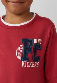 Roter Sweatshirt mit einem weißen Rundhalsausschnitt. Mit einem strukturierten dunkelblauen "FC"-Logo und dem Text "DINO KICKERS", sowie einer Grafik eines Fußballs.