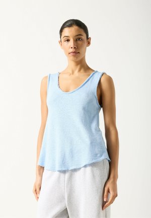 Hellblaues ärmelloses Oberteil mit V-Ausschnitt aus weichem Stoff. Verfügt über ungearbeitete Kanten und einen lockeren Schnitt, kombiniert mit grauen Jogginghosen.