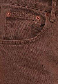 Καφέ ύφασμα τζιν με υφή στην επιφάνεια. Διαθέτει μπροστινή τσέπη με χαλκομανίες και αντίθετη ραφή κατά μήκος των άκρων.