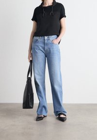 Camiseta corta de manga corta negra con capucha, vaqueros de cintura alta azul claro, bailarinas negras con punta y un bolso tote negro sobre una superficie gris texturizada.