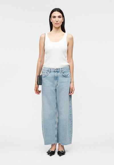 Γυναίκα όρθια, φοράει λευκό αμάνικο τοπ, ανοιχτό μπλε τζιν με φαρδιά μπατζάκια και φθαρμένα τελειώματα, μαύρα μυτερά παπούτσια και μικρή τσάντα χιαστί.