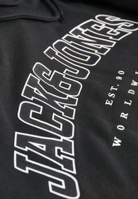 Hoodie noir arborant un grand texte blanc "JACK & JONES", avec des détails fins et le texte "EST. 90 WORLDWIDE" en plus petite police en dessous.