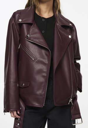 Kobieta ubrana w ciemnoczerwoną, bordową skórzaną kurtkę motocyklową z srebrnymi zamkami i guzikami, na czarnej koszuli i czarnych spodniach.