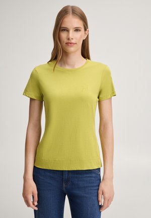 Vrouw met steil lichtbruin haar draagt een aansluitend geelgroen T-shirt en blauwe jeans, staand tegen een effen lichte achtergrond.