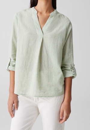 Frau trägt eine hellgrüne, langärmlige Bluse mit V-Ausschnitt und aufgekrempelten Ärmeln, kombiniert mit weißen Hosen.