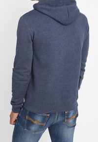 Felpa blu scuro in tessuto strutturato, con cappuccio regolabile tramite coulisse e polsini a coste, abbinata a jeans blu chiaro con cuciture a contrasto sulle tasche.