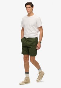 Shorts de algodón verdes con cintura elástica y cordón, combinados con una camiseta blanca de cuello redondo y zapatillas de caña alta beige.
