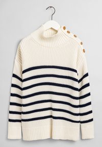 Pull crème à rayures horizontales bleu marine, col montant côtelé, et boutons en bois sur l'épaule. Texture en tricot, coupe décontractée.