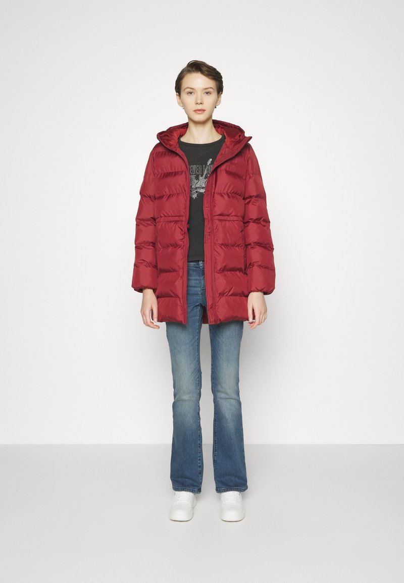 Giacca a vento bordeaux con cappuccio, caratterizzata da un design trapuntato, tasche frontali e chiusura con bottoni, abbinata a jeans a zampa d'elefante blu e scarpe bianche.