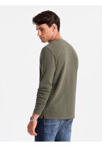 Olivgrünes Langarmshirt mit Rundhalsausschnitt, Seitenschlitzen und einer weichen Textur, kombiniert mit blauen Jeans und einer silbernen Uhr.