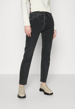 Vrouw die zwarte jeans met hoge taille draagt, een witte trui met crèmekleurige bloemenrand en beige veterschoenen, staand tegen een effen witte achtergrond.