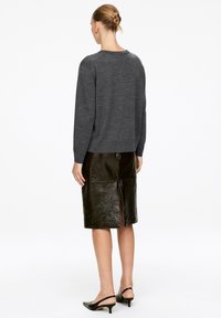 Frau mit einem Dutt, die einen dunkelgrauen Pullover, einen glänzenden schwarzen Lederröck mit hinterem Schlitz und schwarze Slingback-Kitten-Heels trägt, mit dem Rücken zugewandt.