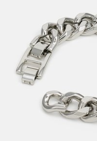 Pulsera de cadena metálica plateada gruesa con un acabado pulido, que presenta eslabones entrelazados y un cierre rectangular.