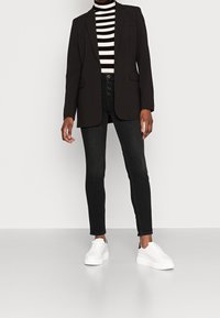 Blazer negro con solapas afiladas, combinado con un jersey de cuello alto a rayas negras y blancas. Jeans oscuros y zapatillas blancas con plataforma completan el conjunto.