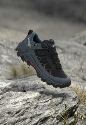 Zwarte en grijze Columbia Titanium waterdichte wandelschoen zwevend boven rotsachtig terrein met gras en kleine witte bloemen.