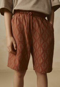 Shorts rouges à motifs avec une taille à cordon, présentant un design de feuilles en blanc. Fabriqués en tissu texturé avec des poches latérales.