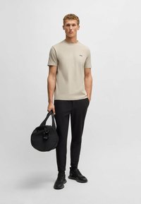 Beige crew-neck t-shirt med sort logo, kombineret med sorte slim-fit bukser og sorte sneakers, holder en rund sort taske med en kort rem.