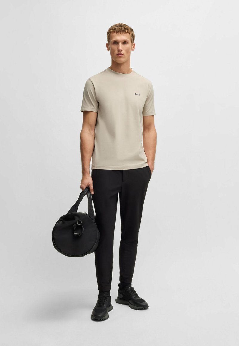 Beige crew-neck t-shirt med sort logo, kombineret med sorte slim-fit bukser og sorte sneakers, holder en rund sort taske med en kort rem.
