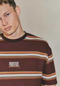 Stribet t-shirt med korte ærmer i mørk maroon, med hvide, orange og brune striber. Teksten "PROVIDENCE" i hvid og orange i midten.