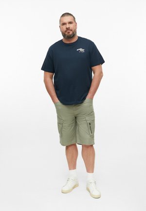 Volwassen man met baard, gekleed in een marineblauw T-shirt, olijfgroene cargoshorts, witte sokken en witte sneakers, staand met handen in zijn zakken.