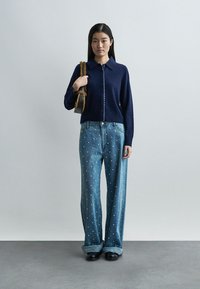 Γυναίκα με μπλε σκούρα ζακέτα με κουμπιά, τζιν παντελόνι φαρδύ με διακοσμήσεις από πέρλες, μαύρα παπούτσια και καφέ υφασμάτινη τσάντα ώμου.