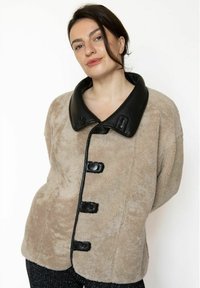 Beige jakke i imiteret pels med stor sort læderkrave, seks sorte knapper foran og en blød, struktureret overflade.