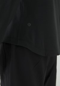 Schwarzes Sportoberteil aus strukturiertem Stoff, mit abgerundetem Saum und kleinem Logo an der Seite, kombiniert mit passenden schwarzen Hosen.