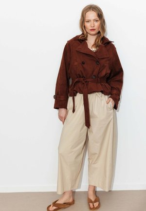 Femme portant une veste en daim marron ceinturée, un pantalon beige à jambes larges et des sandales marron, debout sur un fond blanc avec les mains dans les poches.