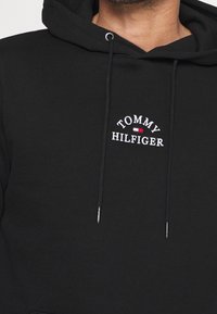 Sudadera negra con "Tommy Hilfiger" bordado en blanco y un pequeño logo de bandera en rojo, blanco y azul en el pecho, con cordones que cuelgan.