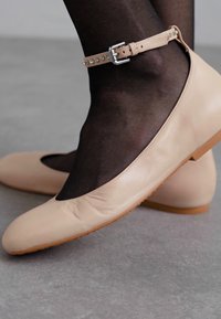 Beige leren ballerina's met enkelbandjes en zilveren gesp, gedragen over zwarte sheer panty’s op een grijze vloer.