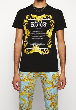 Schwarzes Versace Jeans Couture T-Shirt mit verziertem gelbem Barockrahmen und weißem Text, kombiniert mit blauen Jeans mit passenden gelben Barockmustern.