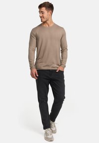 Camicia lunga beige con scollo rotondo, abbinata a jeans scuri e sneakers chiare. Texture liscia, design aderente.