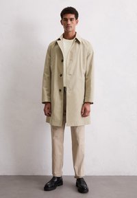 Beige regenjas met grote kraag en donkere knopen, gedragen over een lichtgekleurde shirt en beige broek. Zwarte schoenen maken de outfit compleet.