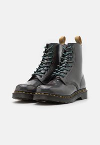 Las botas de tobillo de cuero negro presentan un acabado brillante, cordones azules y verdes, costuras amarillas y tiradores en el cuello. Suela de goma.