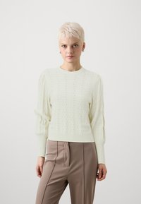 Lys grønn strikkegenser med bølgete mønster, ribbede mansjetter og rund hals, kombinert med beige høy midje bukser.