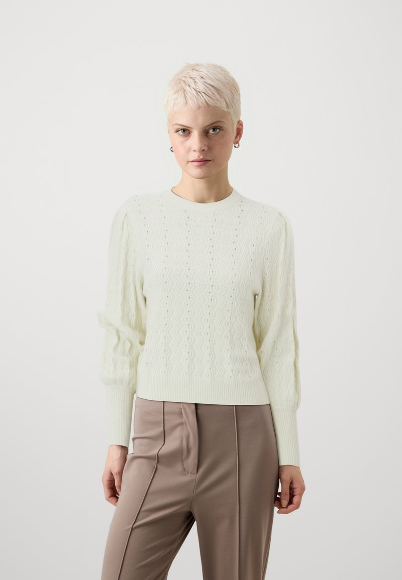 Lys grønn strikkegenser med bølgete mønster, ribbede mansjetter og rund hals, kombinert med beige høy midje bukser.