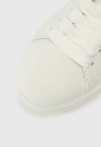 Λευκό δερμάτινο αθλητικό παπούτσι με λεία υφή, στρογγυλεμένη  μύτη και επίπεδα λευκά κορδόνια. Μινιμαλιστικός σχεδιασμός με διακριτική ραφή και λαστιχένια σόλα.