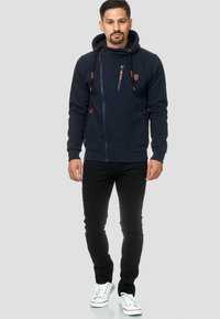 Morska zip-up dukserica s kapuljačom, s bež naglascima, prednjim zip džepom i špagama. Uparena s crnim trapericama i bijelim tenisicama.