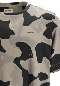 Camiseta de manga corta tipo crewneck en beige con patrón de camuflaje en negro y gris; presenta un logo de marca y costuras en contraste.