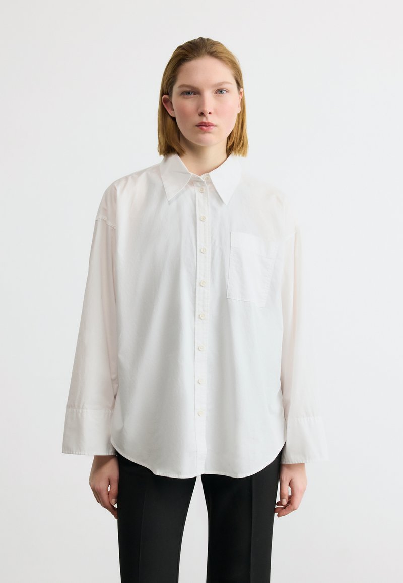 Νεαρή γυναίκα φοράει λευκό πουκάμισο μεγάλου μεγέθους με κουμπιά και μαύρο παντελόνι, στέκεται μπροστά σε απλό λευκό φόντο, κοιτάζοντας προς τα εμπρός.