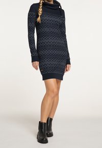 Laivastonsininen neulemekko, jossa on kaulakaulus ja pitkät hihat. Mekko on varustettu valkoisilla geometrisilla kuvioilla, ribbaiduilla hihansuilla ja logotuksessa helmassa.