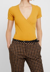 Senapsgul V-halsad ribbad bodysuit med korta ärmar, tillsammans med högmidjade svarta och orange rutiga byxor och ett svart bälte.