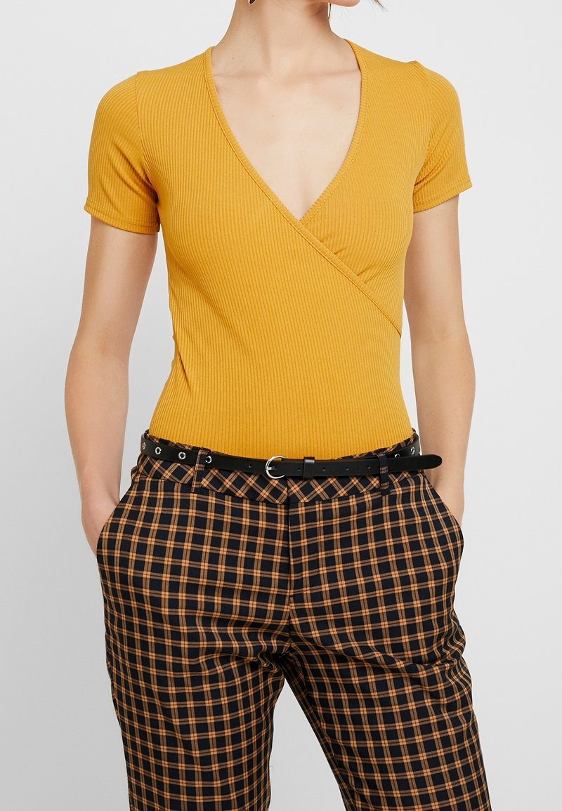 Senapsgul V-halsad ribbad bodysuit med korta ärmar, tillsammans med högmidjade svarta och orange rutiga byxor och ett svart bälte.