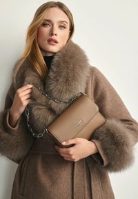 Kobieta w brązowym płaszczu z dużym futrzanym kołnierzem i mankietami, trzymająca torebkę ze skóry taupe z srebrnym łańcuchem i małym metalowym logo.