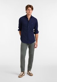 Camisa de botones azul marino, con mangas largas arremangadas, combinada con pantalones verde oliva y zapatillas con cordones crema y detalles marrones.