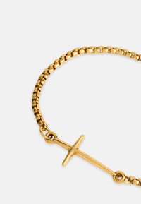 Zlatý náramok s textúrovaným reťazcovým dizajnom, zdobený jednoduchým krížovým príveskom. Kov má lesklý povrch a odráža svetlo.